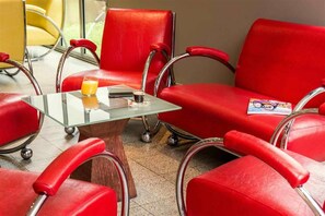 A seating area with red chairs and a glass table, a smartphone, a glass of orange juice, and a magazine. - Ibis Villepinte Parc Expos (Villepinte)