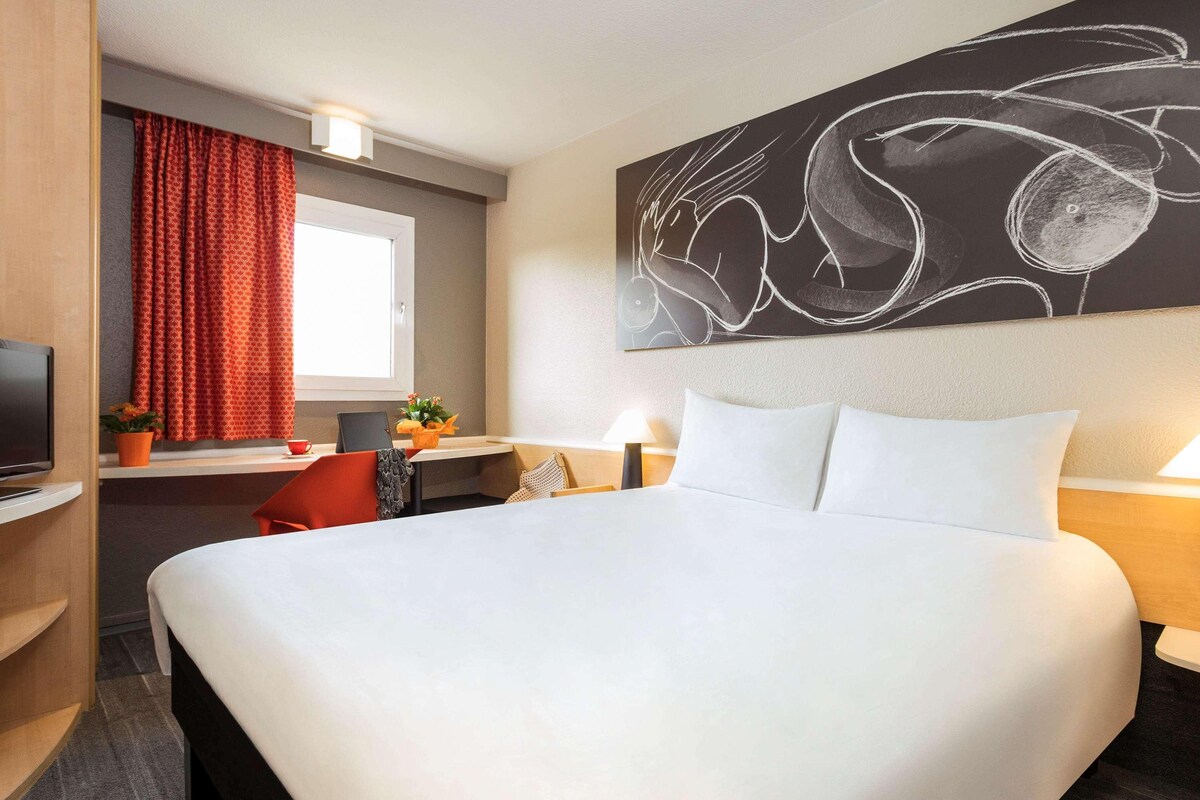 A hotel room with a large bed, a desk with a computer, a TV, and a painting on the wall.