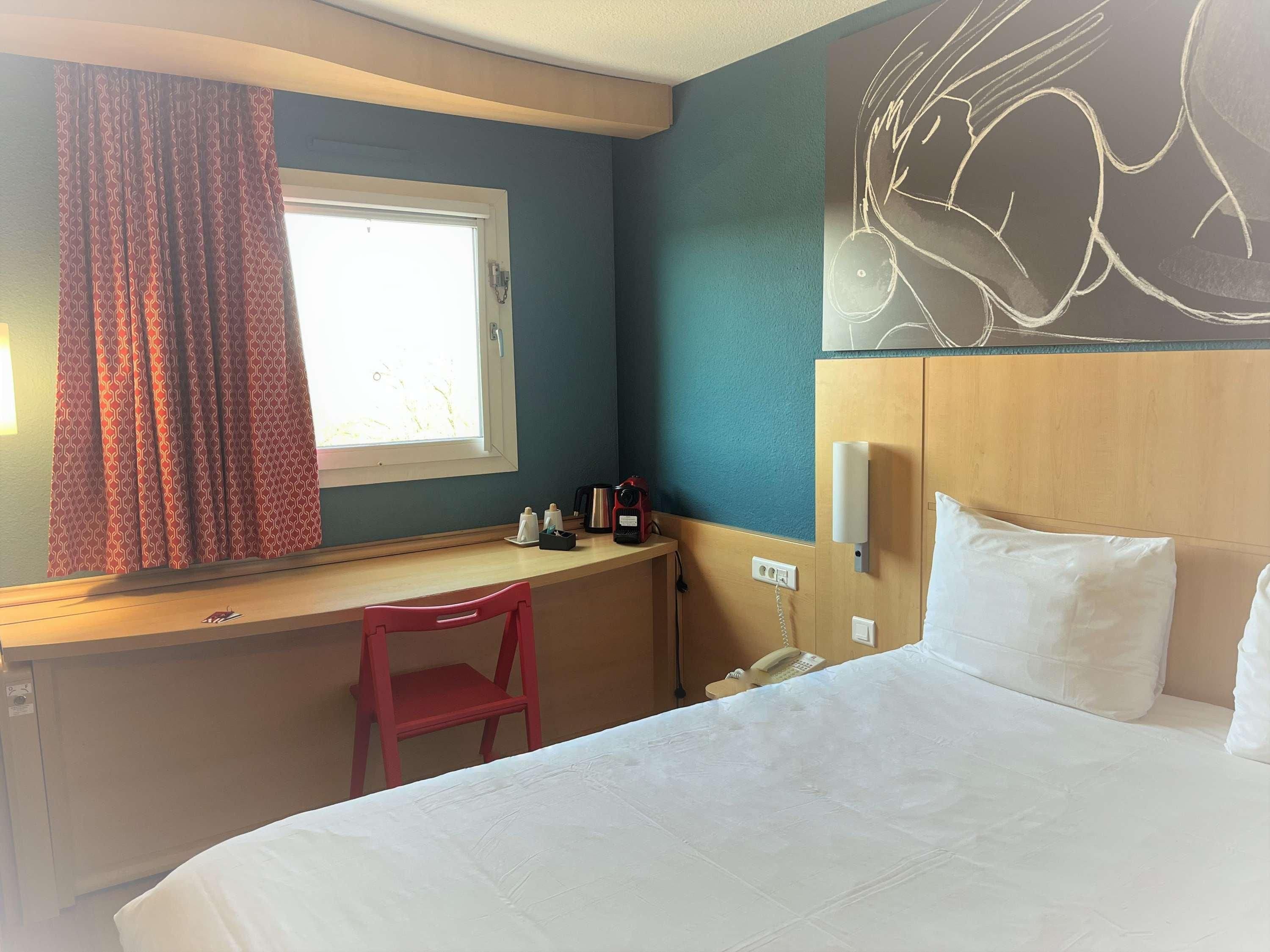 Una habitación de hotel con cama, escritorio y silla, ventana con cortinas, un reloj de pared y teléfono instalado en la pared.