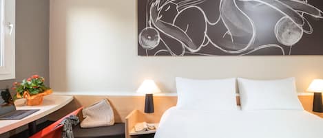 A hotel room with a bed, a desk with a laptop, a chair, a nightstand with a lamp, and a wall art piece.