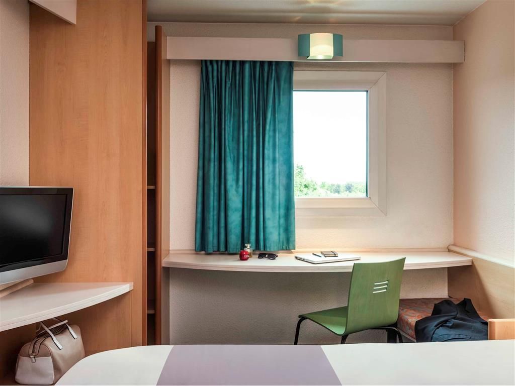 Une chambre d’hôtel comprenant un lit, un bureau avec une chaise, une télévision, une fenêtre avec des rideaux et une petite table avec une lampe.