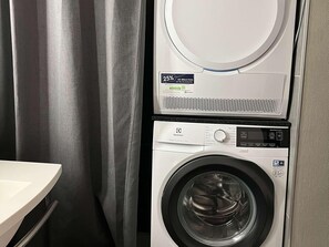 A front-loading washing machine with a digital control panel, a dryer above it, and a curtain to the left. - Ibis Villepinte Parc Expos (Villepinte)
