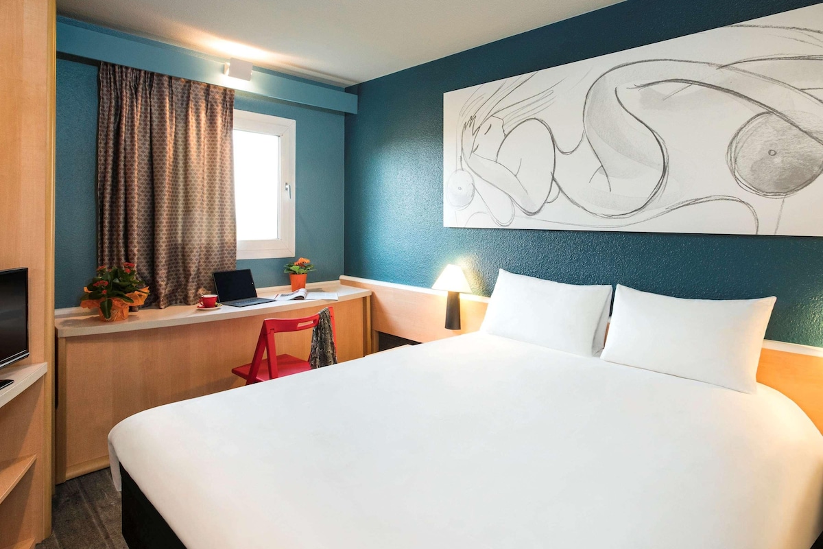 A hotel room with a bed, desk, chair, TV, and artwork on the wall.