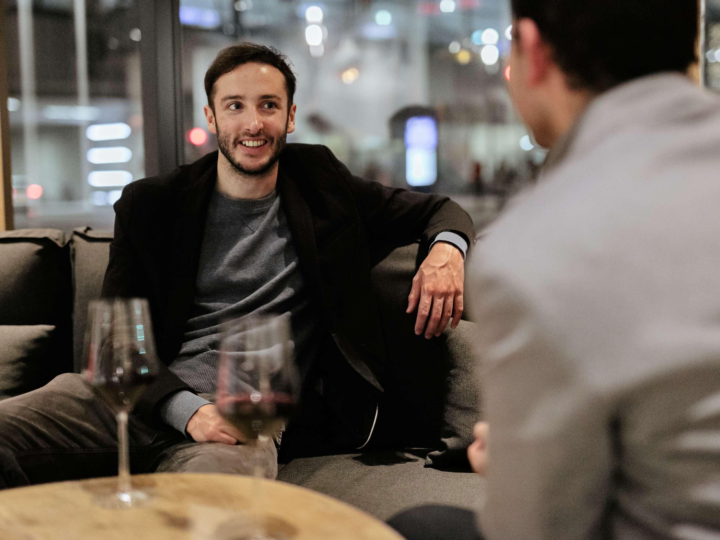 A person sitting on a couch with a glass of wine, another person facing them, and a blurred background suggesting an indoor setting.
