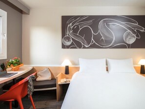 Una habitación de hotel con cama, escritorio, silla, mesita de noche, lámpara y una obra de arte en la pared.