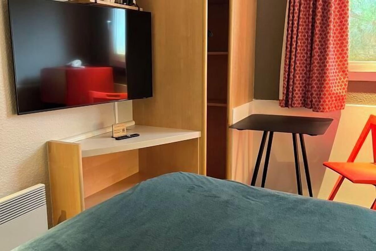 A room with a bed, a TV mounted on the wall, a desk, a chair, and a window with curtains.