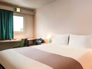 A hotel room with a large bed, a desk with a chair, a window with curtains, and a nightstand with a lamp. - Ibis Villepinte Parc Expos (Villepinte)