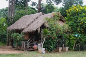 Een landelijke, rieten hut met een veranda, omringd door weelderig groen en bloeiende planten.