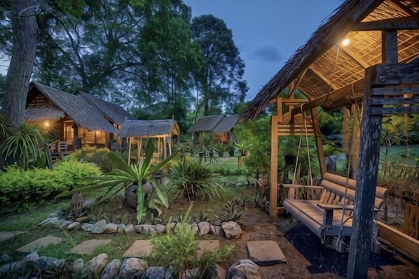 A rustic lodge with thatched-roof cabins, a wooden swing bridge, and a well-maintained garden.