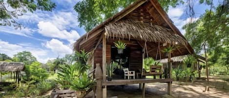Una cabaña de madera con techo de paja, rodeada de follaje y mobiliario de exterior.