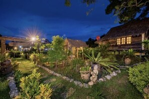 Ein gut beleuchteter Garten mit einem strohgedeckten Gebäude, umgeben von verschiedenen Pflanzen und Steinen.