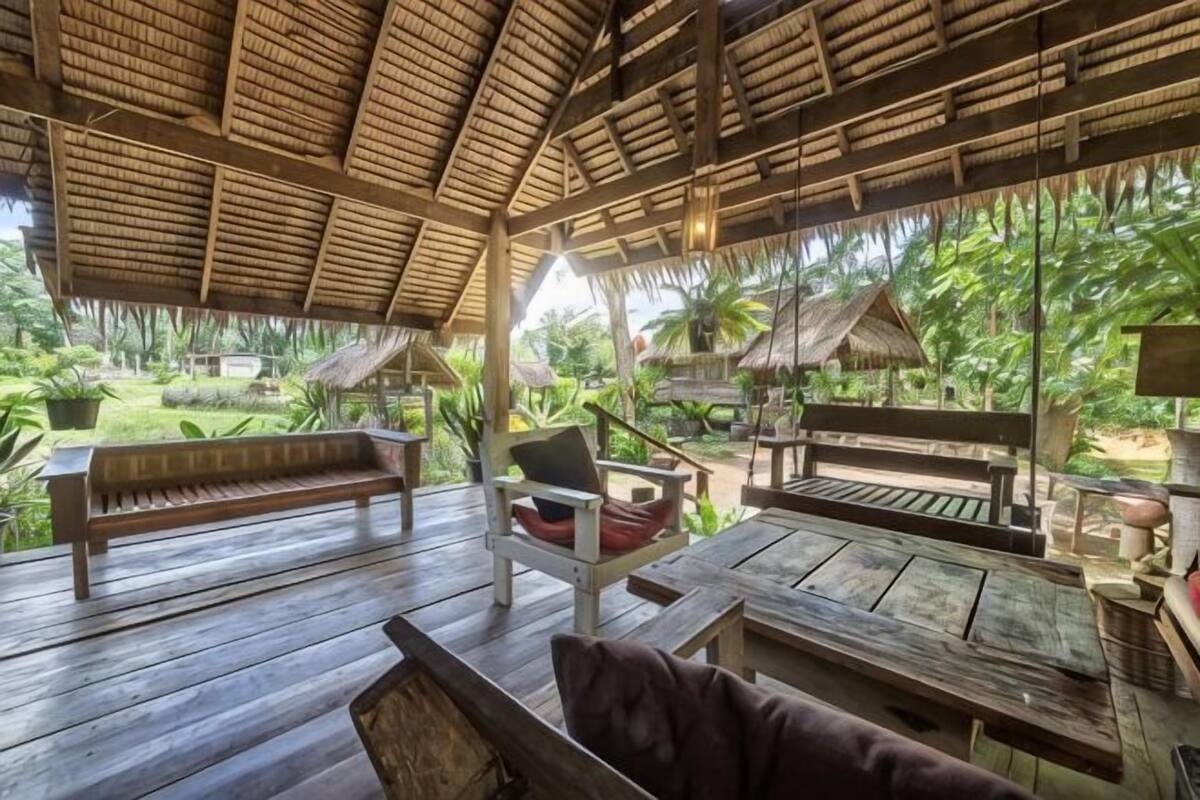 Une terrasse extérieure en bois, dotée de bancs et de tables, entourée d’une végétation luxuriante et surmontée d’une structure à toit de chaume.