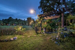 Een tuinpad dat leidt naar een houten pergola met een bord, omringd door groen en een hek.