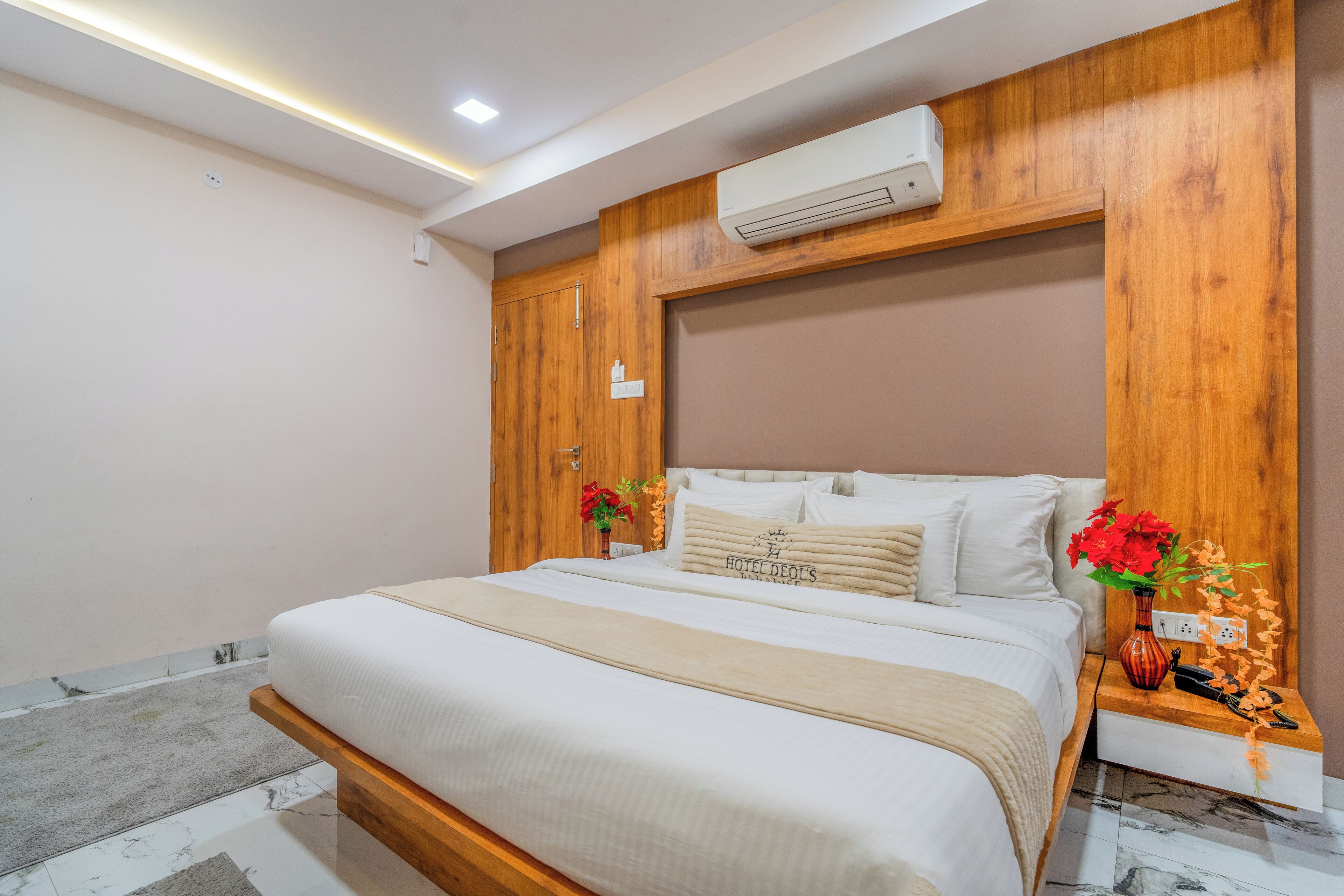 A neatly made bed with a headboard, a bedside table with a vase of flowers, and an air conditioning unit mounted on the wall.