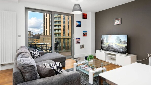 Een moderne woonkamer met een grijze bank, een glazen salontafel, een flatscreen televisie en stadszicht door grote ramen.