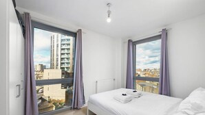 Een moderne hotelkamer met een groot bed, twee ramen met gordijnen, uitzicht op de stad en een plafondlamp.