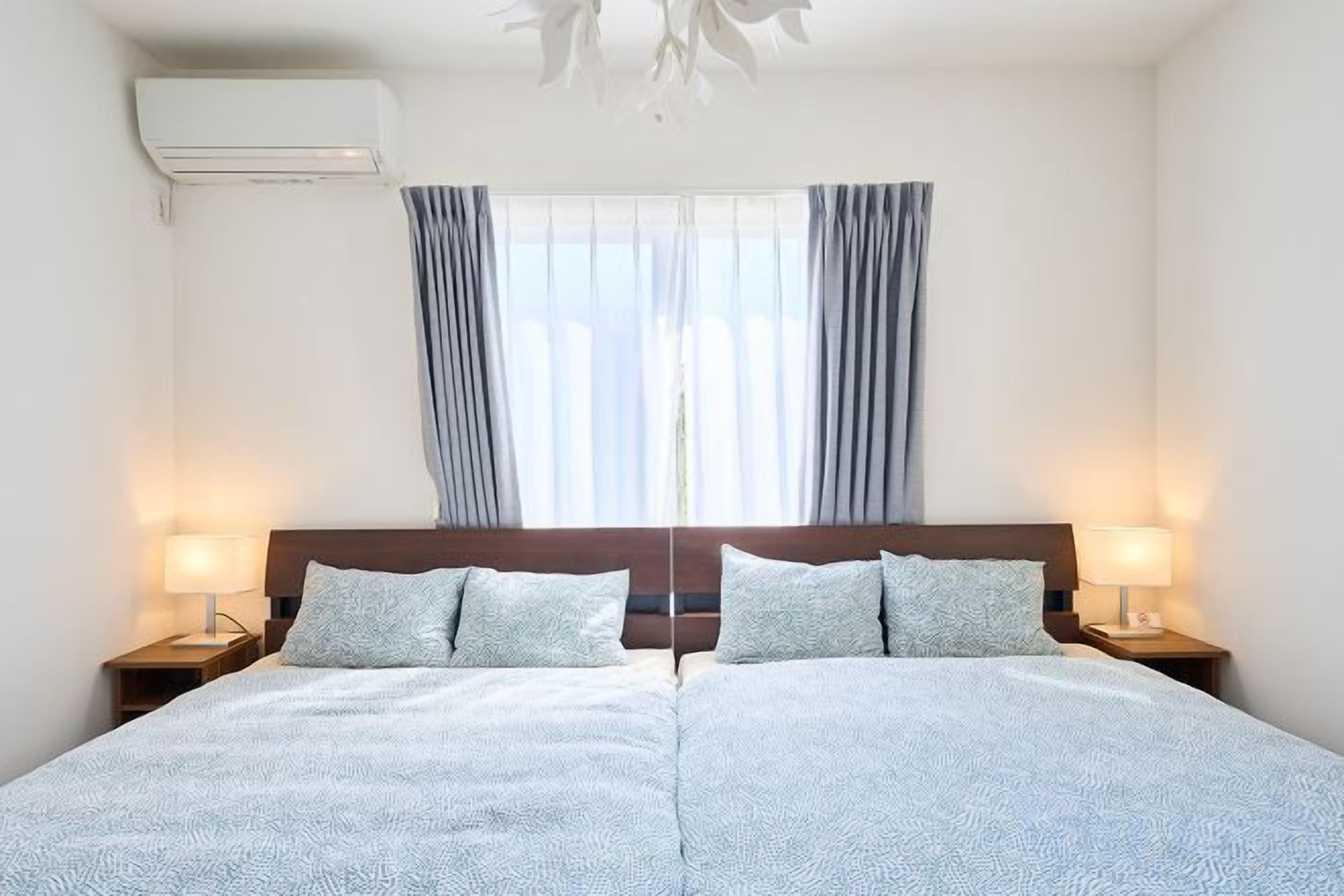 A double bed with a headboard, two bedside tables with lamps, a window with curtains, and an air conditioning unit.