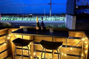 Un espace bar extérieur avec une table en bois et deux tabourets de bar, deux bouteilles de champagne et des verres.