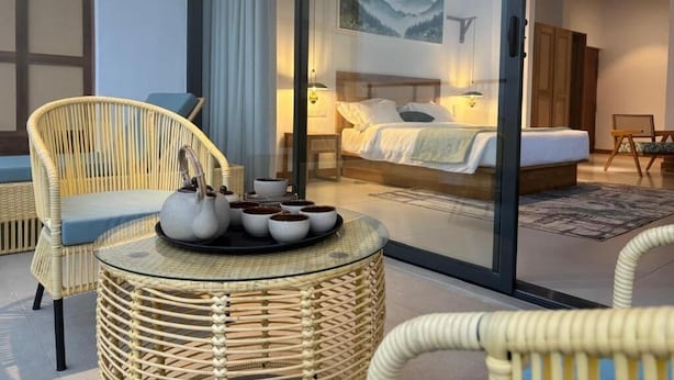 Een moderne hotelkamer met een glazen scheidingswand, een bed, een rieten stoel en een klein tafeltje met theepotten.