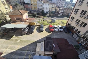 An aerial view of a parking lot with several cars, a small building with a tiled roof, and surrounding multi-story buildings.