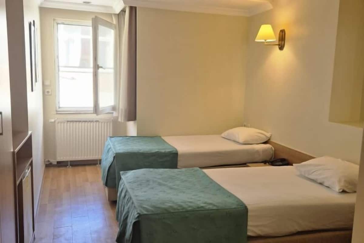 A hotel room with two beds, a window with curtains, a radiator, and a wall-mounted lamp.