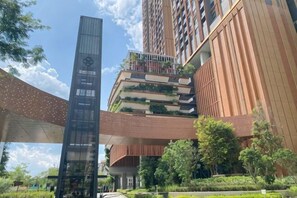 Un edificio moderno de gran altura con una estructura vertical destacada, un jardín en la azotea y un puente peatonal.