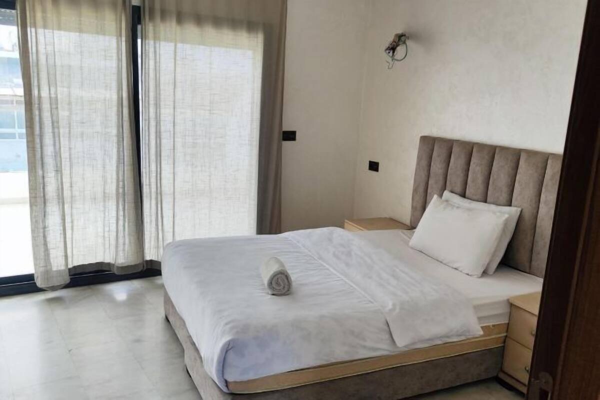 Habitación de hotel con cama, mesita de noche, lámpara de pared y vista a través de una ventana con cortinas.