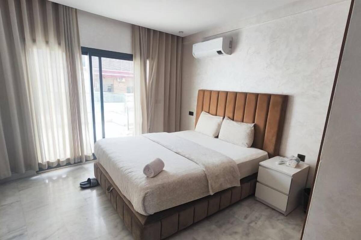 Habitación de hotel moderna con una cama grande, una mesita de noche, aire acondicionado instalado en la pared y vistas amplias a través de un ventanal.