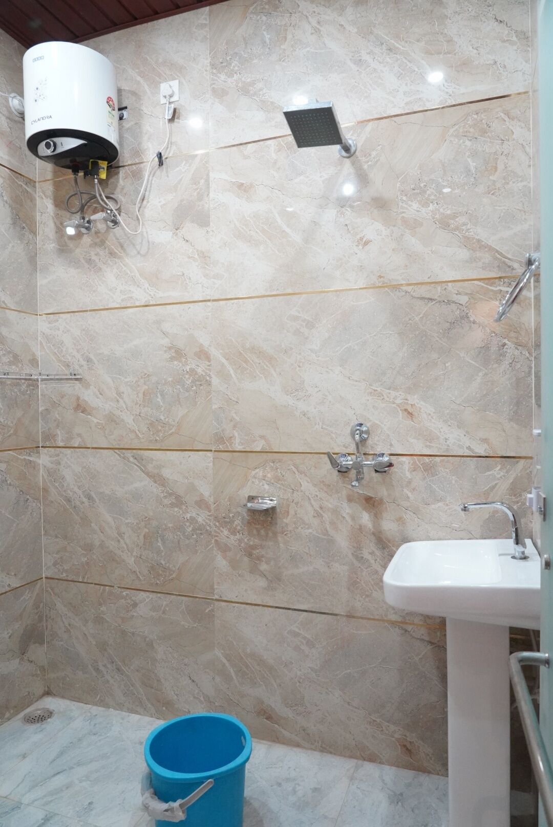 A bathroom with marble walls, a white sink, a shower with a handheld showerhead, and a blue bucket.