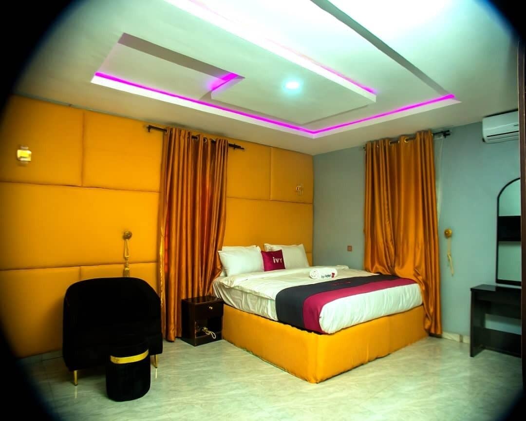 A modern hotel room with a large bed, orange curtains, and a bright ceiling with LED lighting.