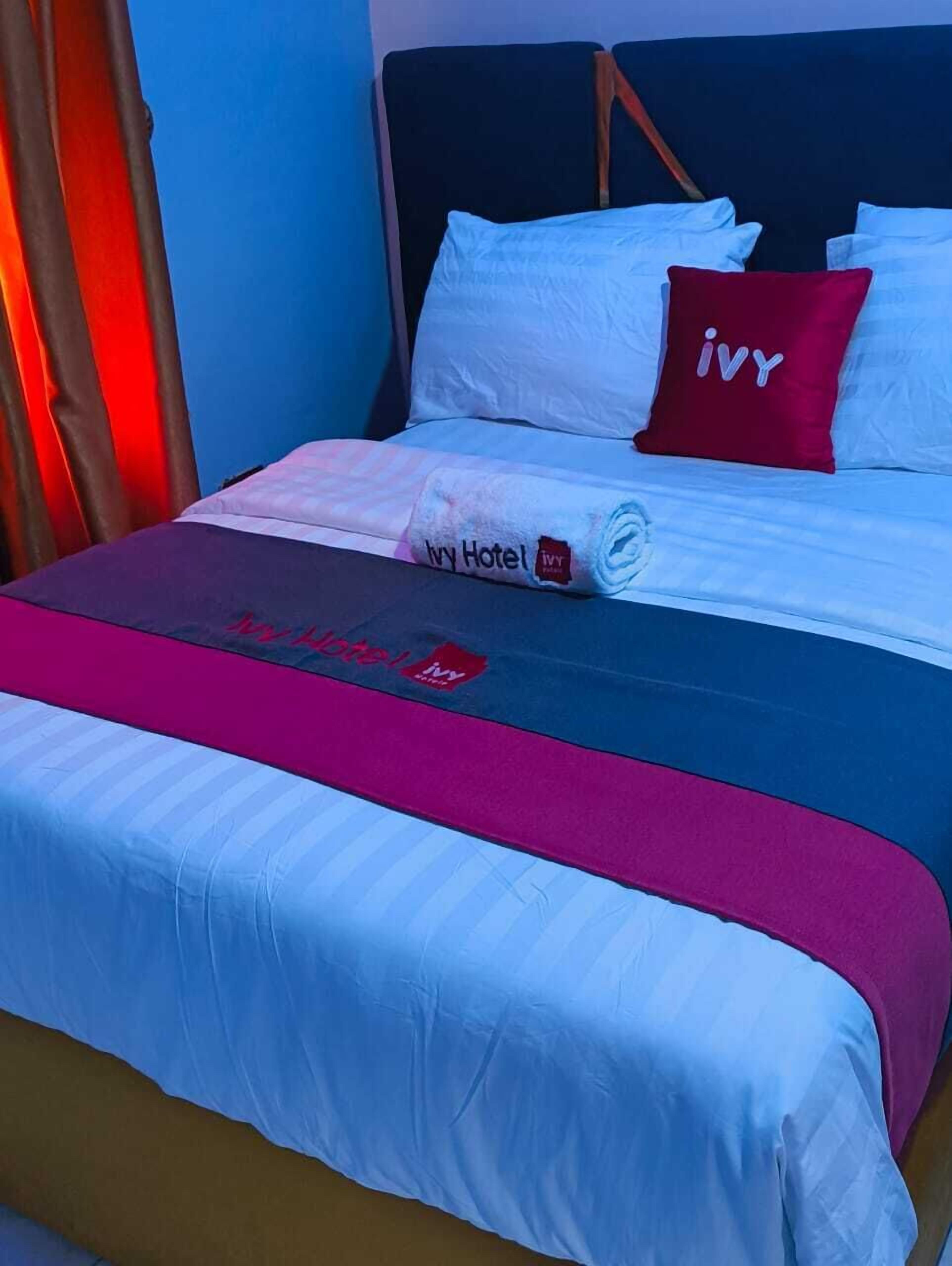 A hotel room with a bed featuring a blue and white bedspread, a red pillow with the hotel logo, and a rolled-up towel on the bed.