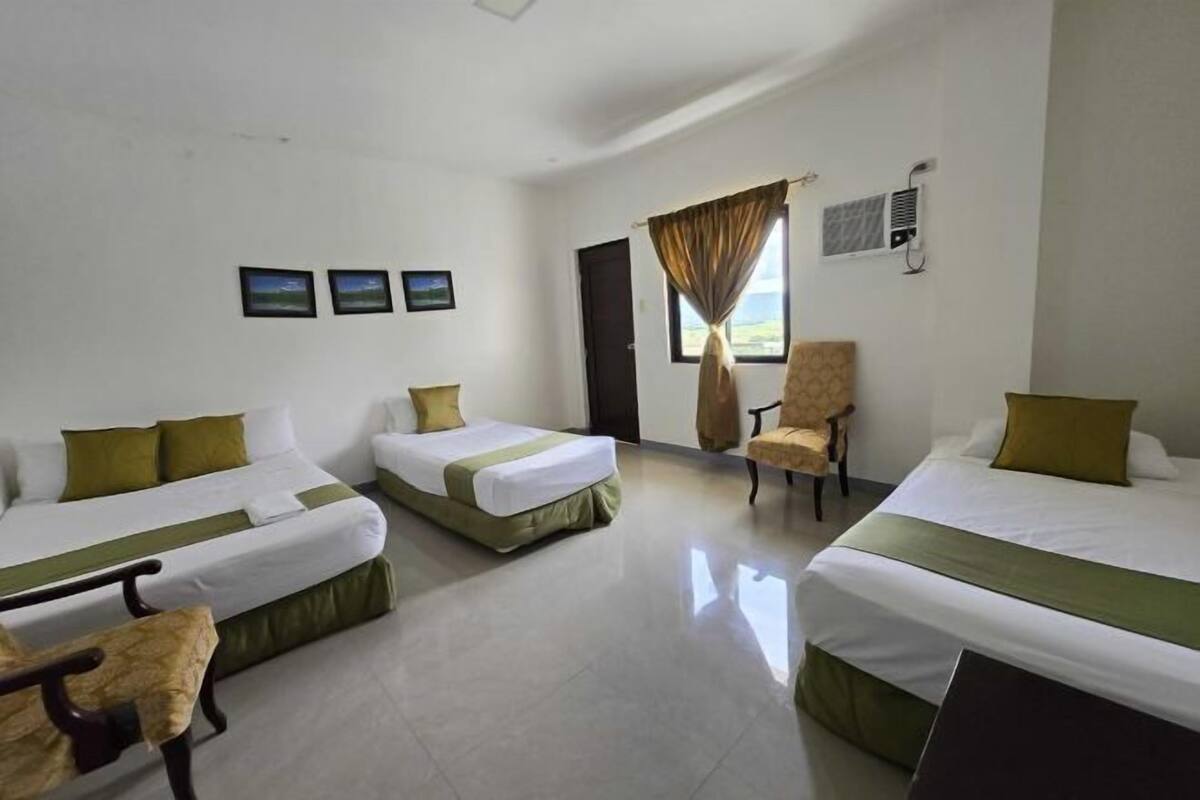 Habitación de hotel con tres camas, una silla, un ventanal con cortinas y un aire acondicionado instalado en la pared.
