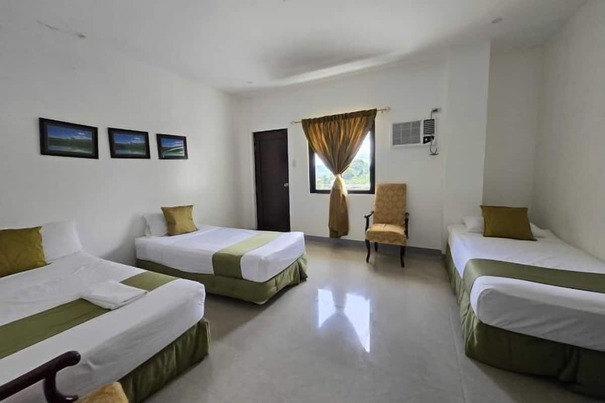 Habitación de hotel con tres camas individuales, una silla, un ventanal con cortinas y tres cuadros en la pared.