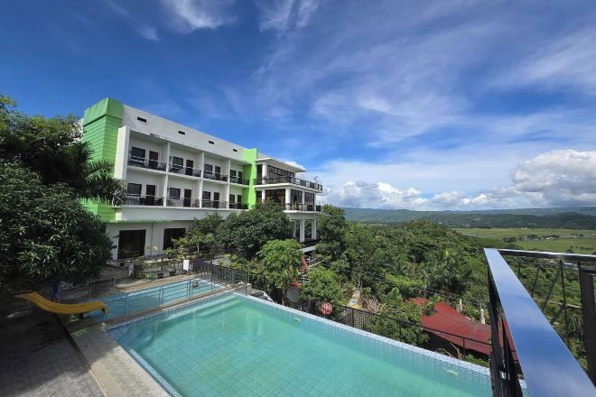A hotel with a pool, surrounded by lush greenery and a view of distant hills.