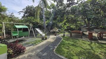 Un parque infantil con tobogán y columpios, rodeado de vegetación y un sendero de grava.