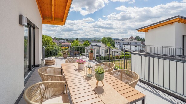 Un balcón con mesa de madera y sillas de mimbre, con vistas a una zona residencial con casas y vegetación.
