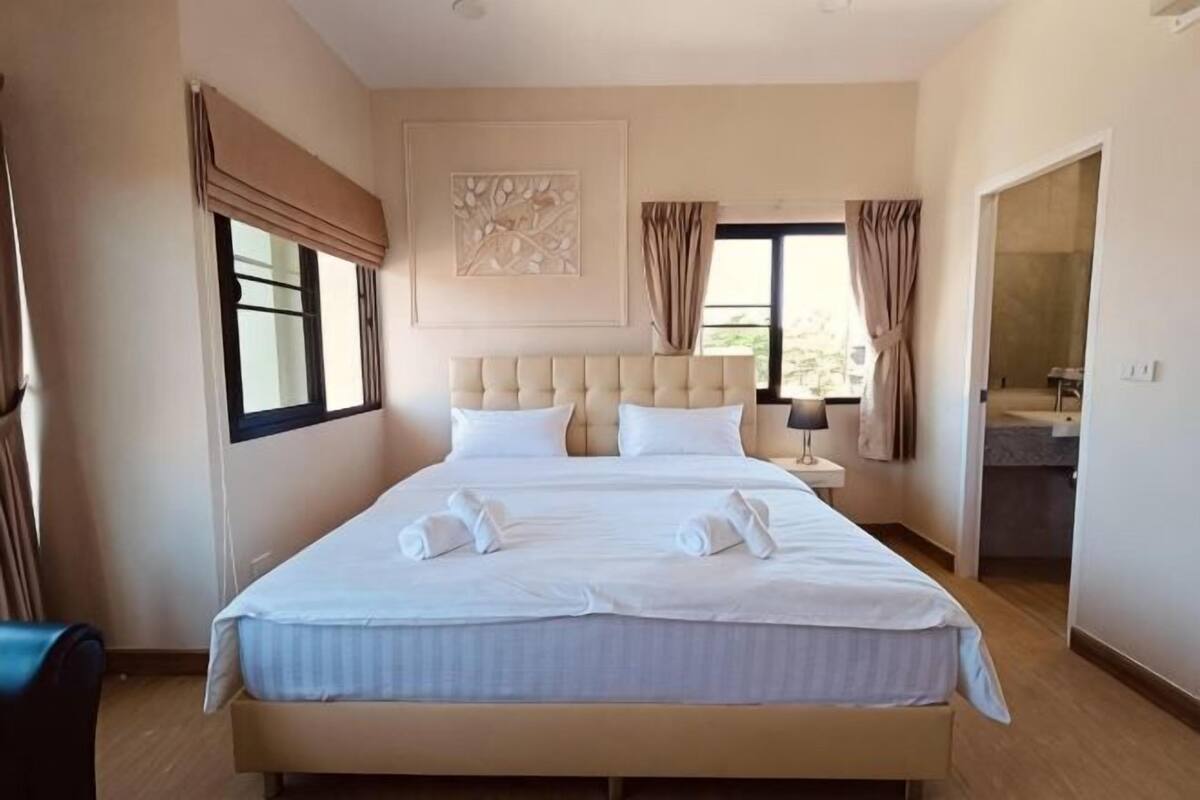 A large bed with a headboard, a bedside table, a window with curtains, and a bathroom visible through an open door.
