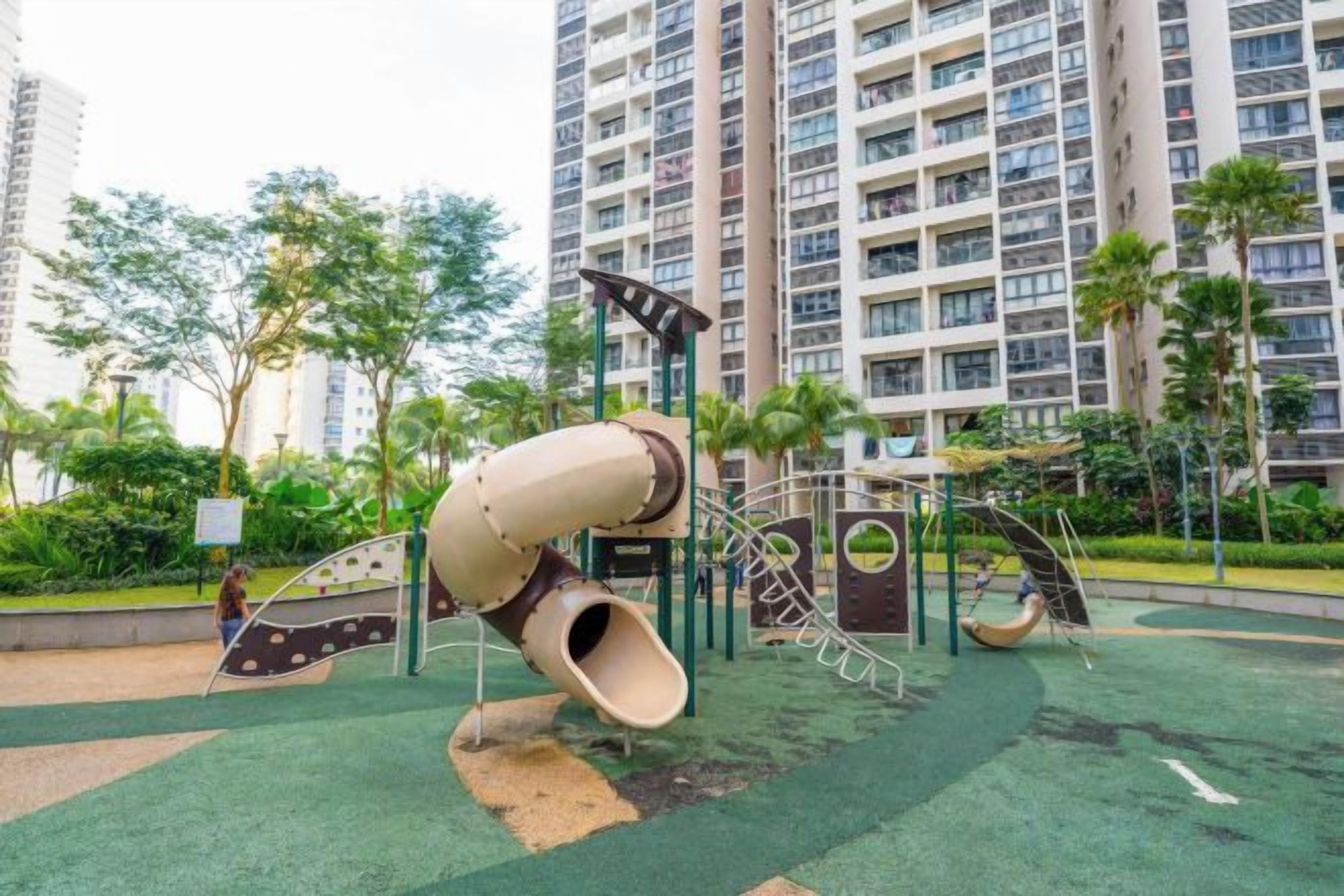 A playground with a spiral slide and climbing structures, surrounded by greenery and tall residential buildings.
