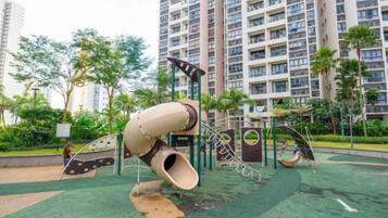 A playground with a spiral slide and climbing structures, surrounded by greenery and tall residential buildings.