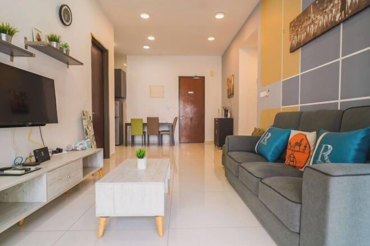 A modern living room with a flat-screen TV, a coffee table, a sofa with decorative pillows, and a dining area in the background.