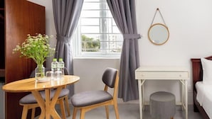 A room with a wooden dining table, two chairs, a small stool, a white desk, a round mirror, and a bed with white bedding. - Luxury Vuon Lai Hotel (Ho Chi Minh City)