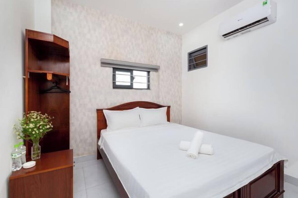 Un lit bien fait, avec du linge de lit blanc et une serviette pliée, une armoire en bois, une petite fenêtre et un appareil de climatisation fixé au mur.