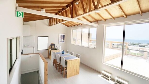 A modern interior with a wooden ceiling, a dining area with a long table and chairs, large windows with a view, and a small kitchenette. - and SEA The Terrace (Kamogawa)