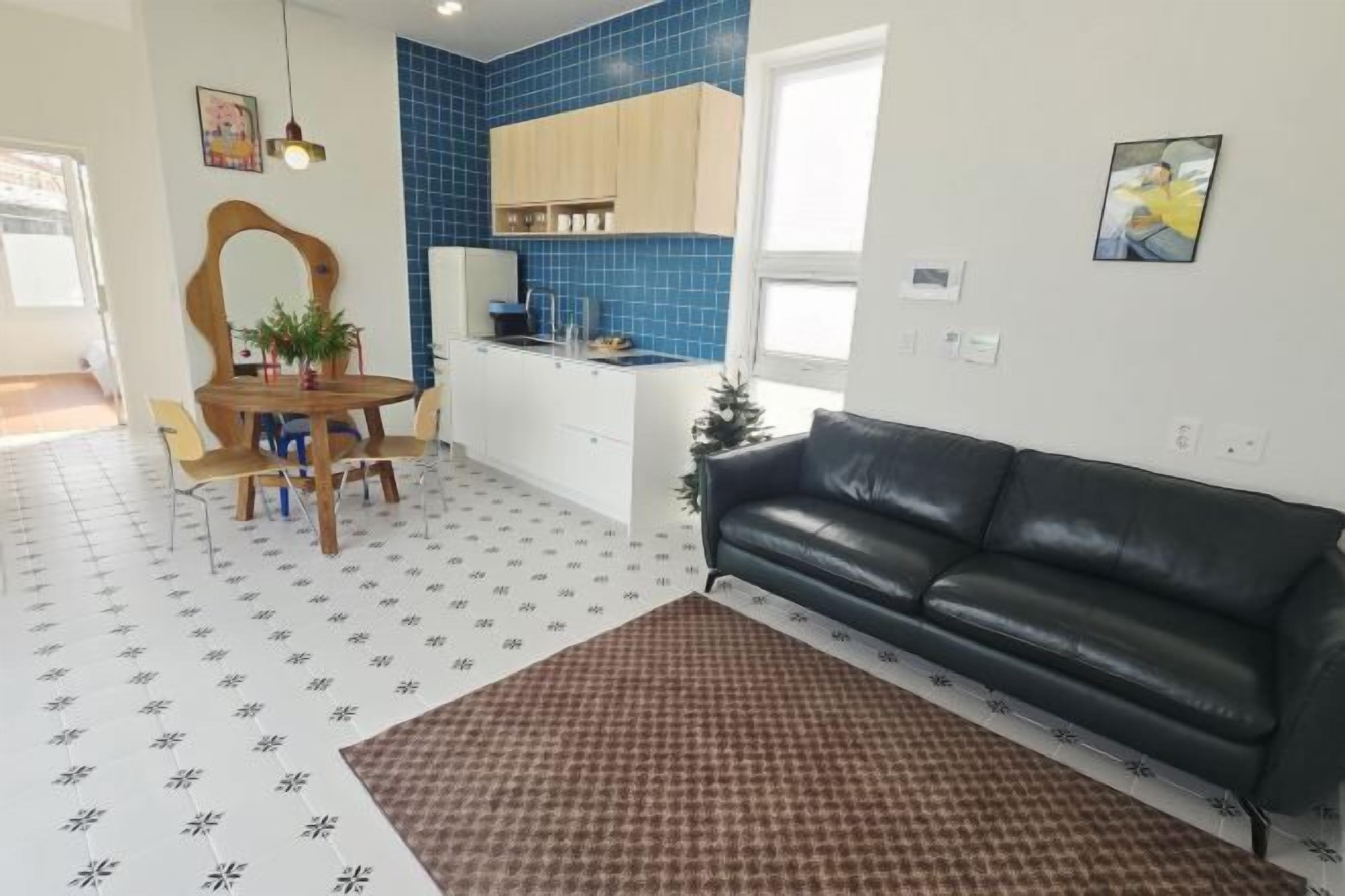 A modern living room with a black leather sofa, a dining table with chairs, a kitchen area with blue tiles, and a patterned rug.