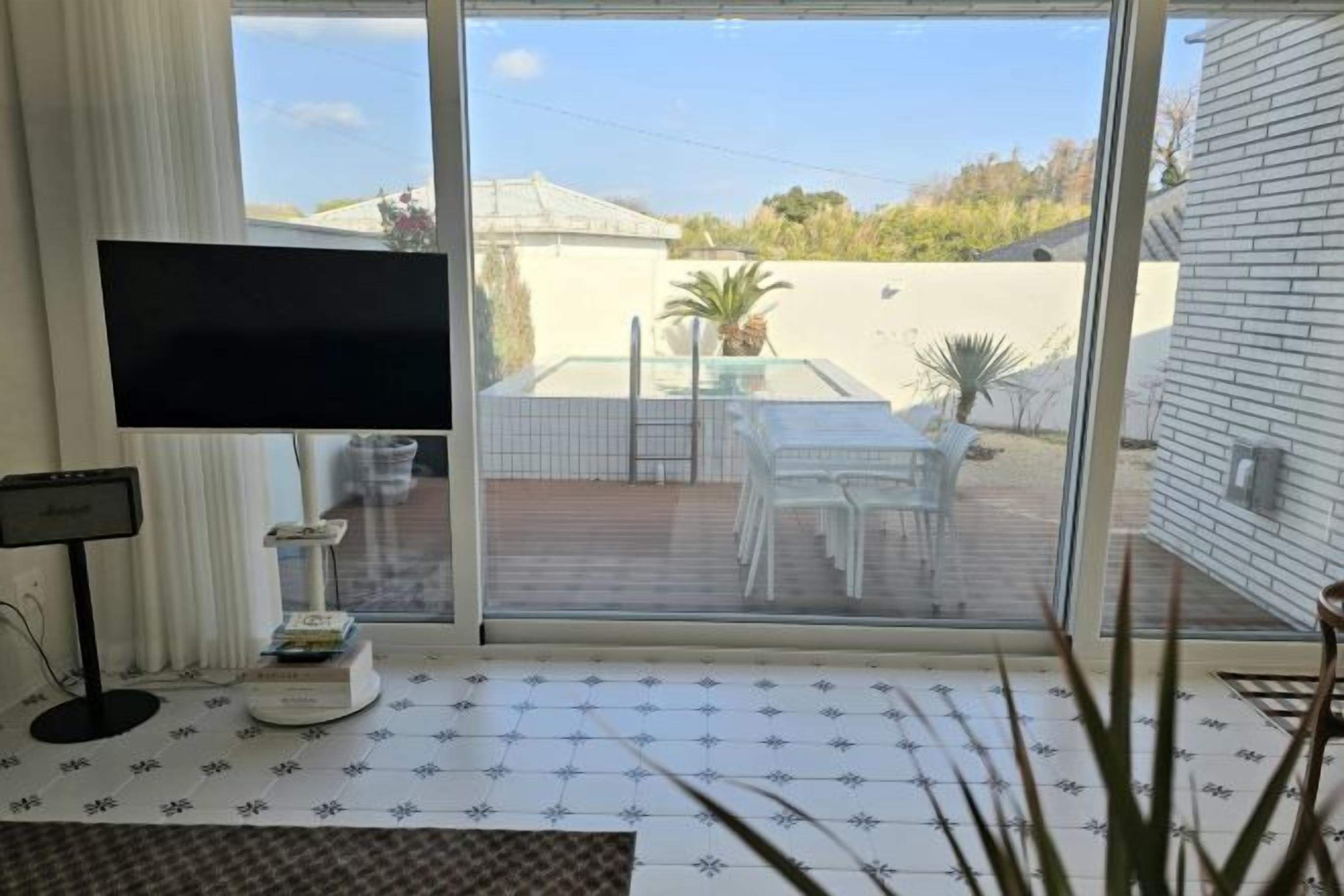 Een kamer met een aan de muur bevestigde televisie, een klein tafeltje met boeken, een glazen schuifdeur naar een buiten terras met een tafel en stoelen, en een plant in een pot.