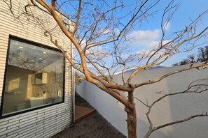 Un edificio moderno con un gran ventanal que refleja el interior de un baño, un árbol en primer plano y un techo inclinado.