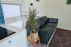 Un espacio de estar con un sofá, una planta en maceta, una mesita y una ventana con persianas.