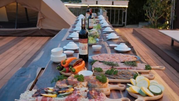 A long wooden table set for a meal with various meats, cheeses, and vegetables, a bottle of wine, and a tent in the background. - Mountain Nest Villa Dalat - BBQ, Cloud Hunting, Camping (Lac Duong District)
