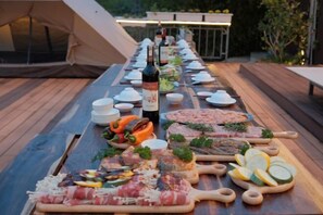 Una larga mesa de madera dispuesta para una comida con carnes, quesos y verduras variados, una botella de vino, y una tienda de campaña al fondo.