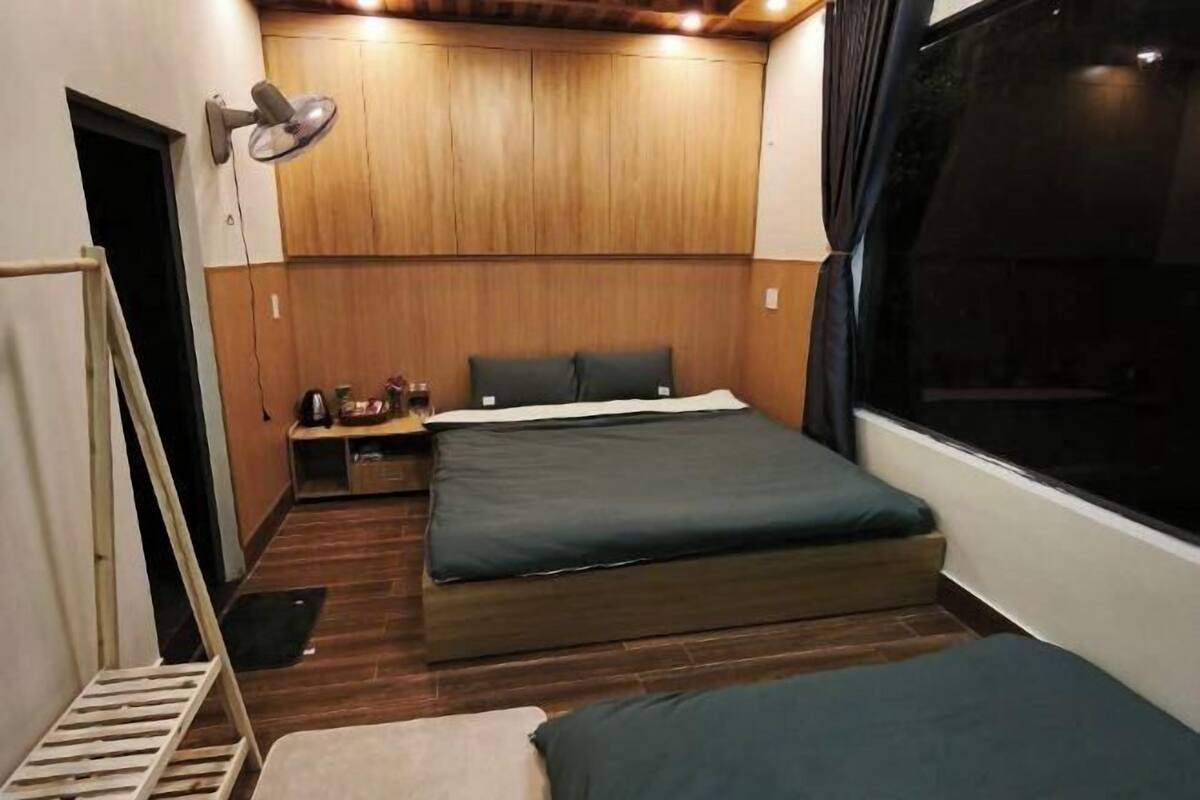 A compact bedroom with a wooden headboard, a bed with dark bedding, a bedside table with a lamp, and a window with curtains.